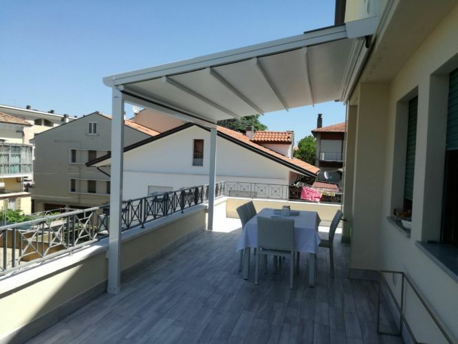 Terrazza coperta sul tetto con tavolo e sedie bianchi, con vista sugli edifici circostanti sotto un cielo limpido.