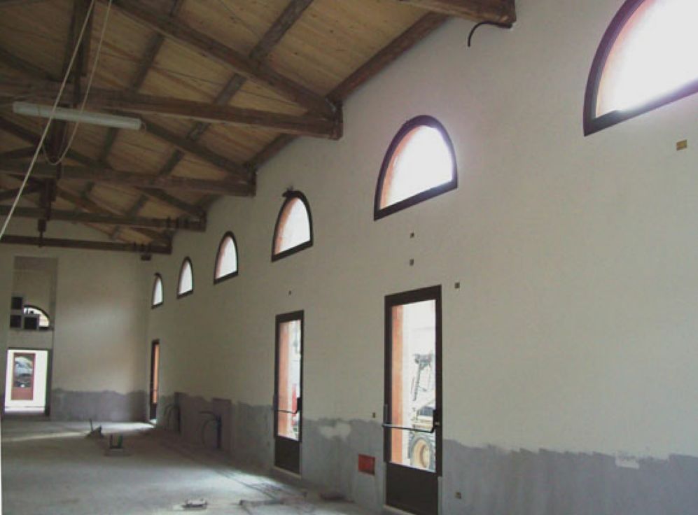 Sala vuota con pareti bianche, finestre ad arco, porte scure e travi di legno a vista sul soffitto.