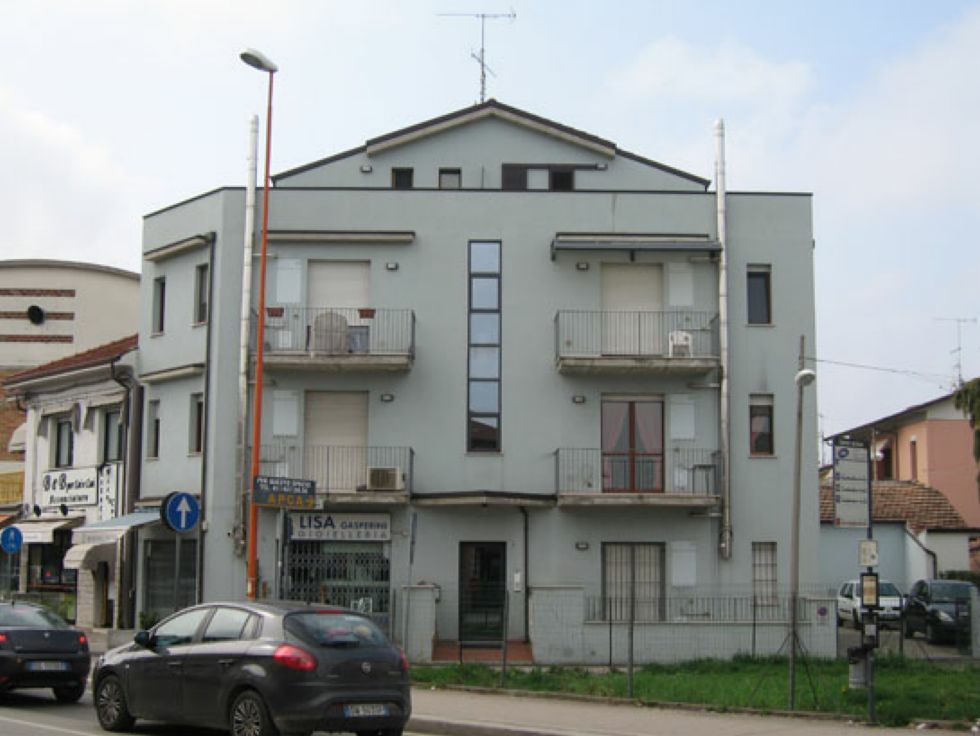 Edificio residenziale di tre piani con balconi, situato all'angolo di una strada, con auto parcheggiate di fronte.