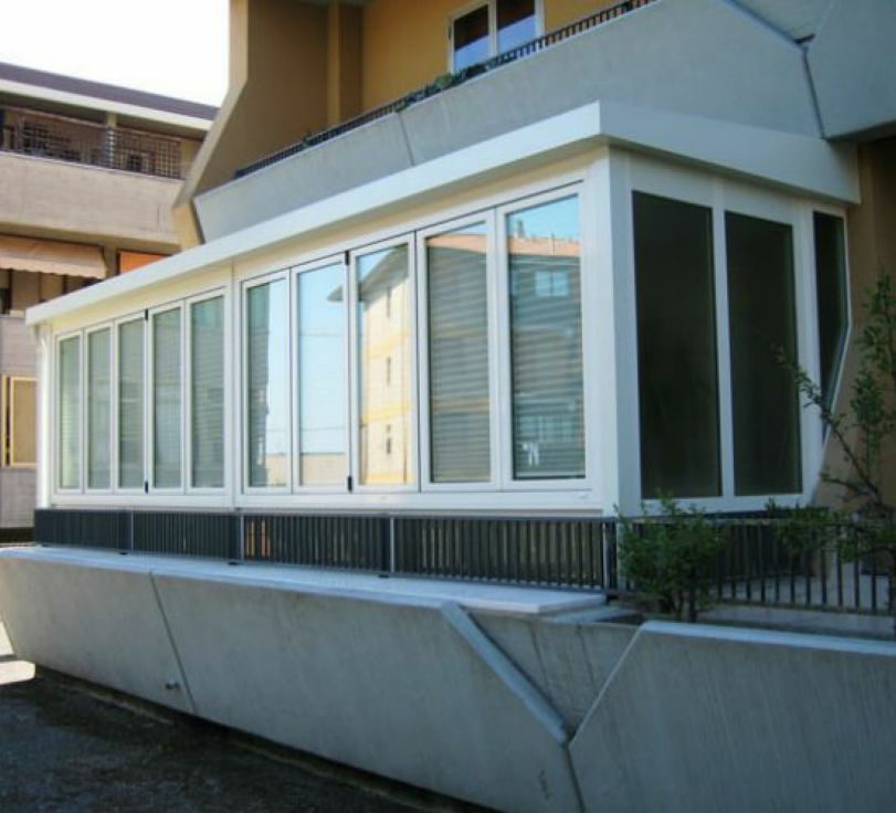Veranda bianca chiusa con ampie finestre accanto a un edificio beige e a un muro di cemento inclinato.