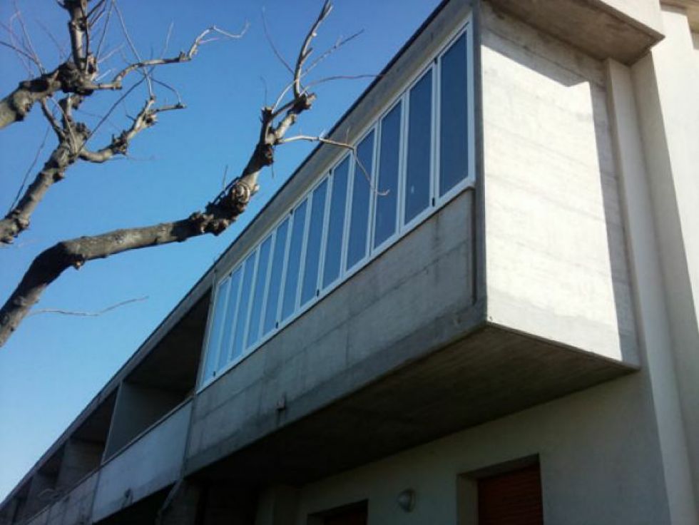 Esterno di un edificio moderno con balcone in cemento e finestre dai colori blu contro un cielo limpido.