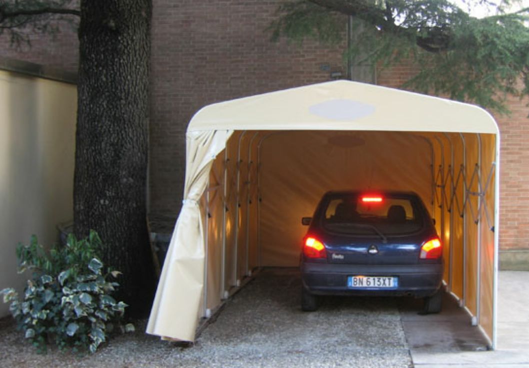 Auto blu parcheggiata all'interno di una tettoia beige con le luci posteriori accese, accanto a un albero e a un vialetto di ghiaia.