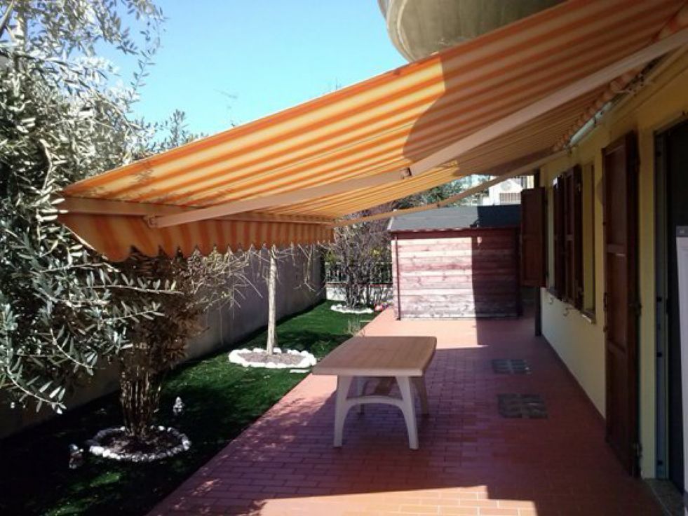 Patio ombreggiato con tenda a strisce arancioni, pavimento in mattoni, tavolo e muro della casa accanto a un giardino.