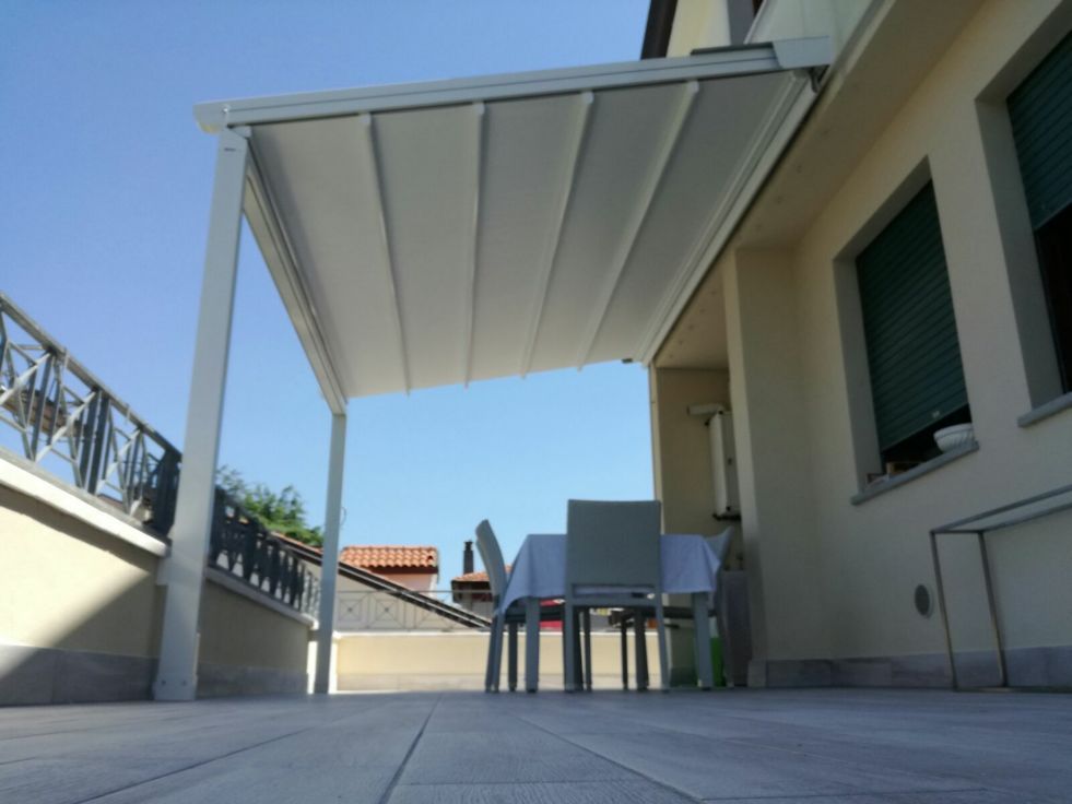 Patio con tenda da sole bianca, tavolo e sedie accanto a una casa sotto un cielo azzurro limpido.