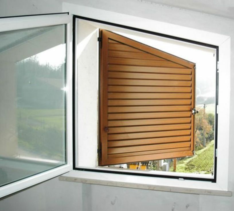 Finestra aperta con pannello di persiana in legno marrone incernierato verso l'esterno, all'interno di una stanza bianca.