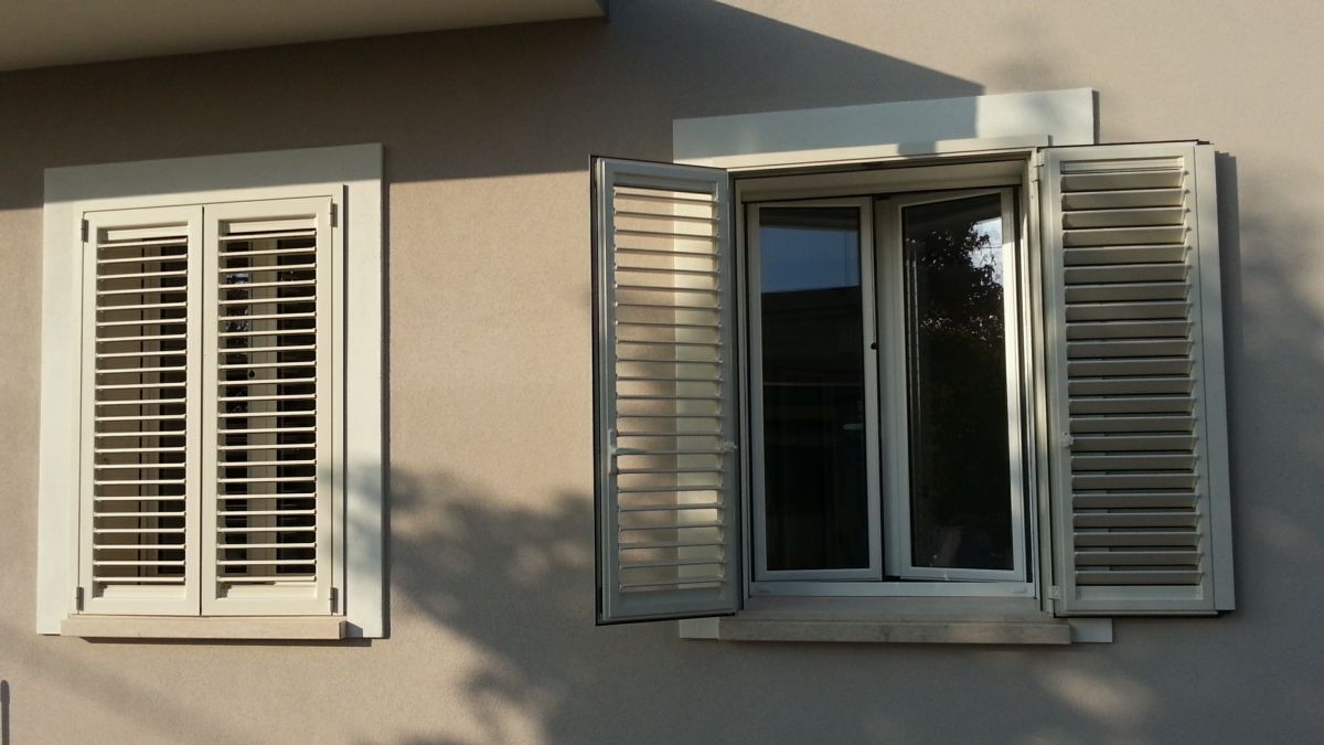 Due finestre con persiane bianche su una facciata di casa beige, una aperta e una chiusa.