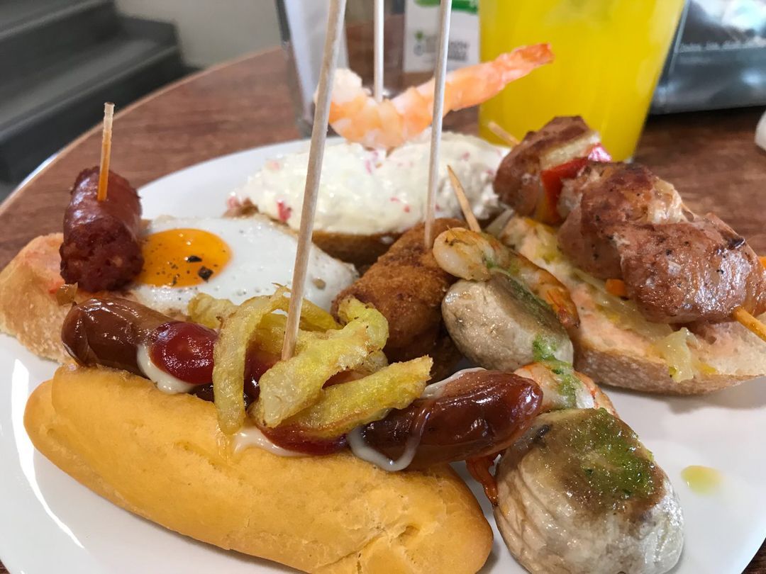 Assorted tapas on skewers and bread, with sausages, shrimp, and sauces on a white plate