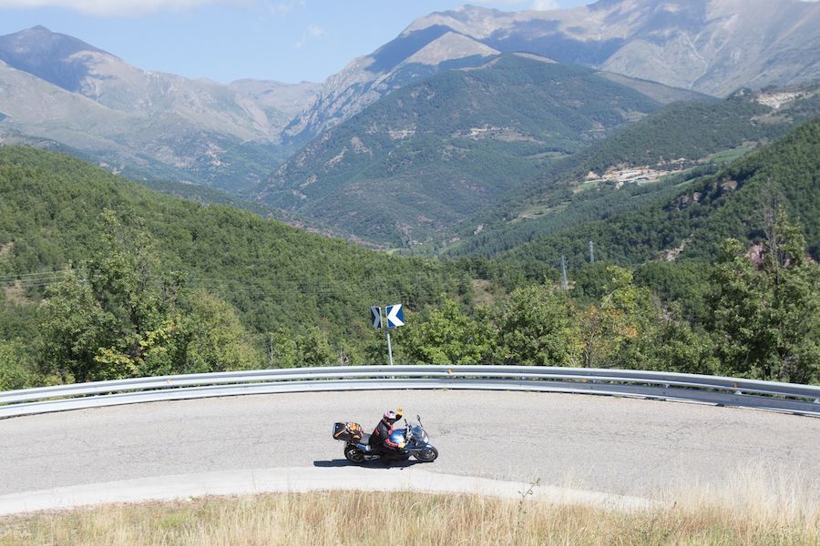 Motorcycle leaning into a sharp mountain road curve with forested hills and peaks in the background