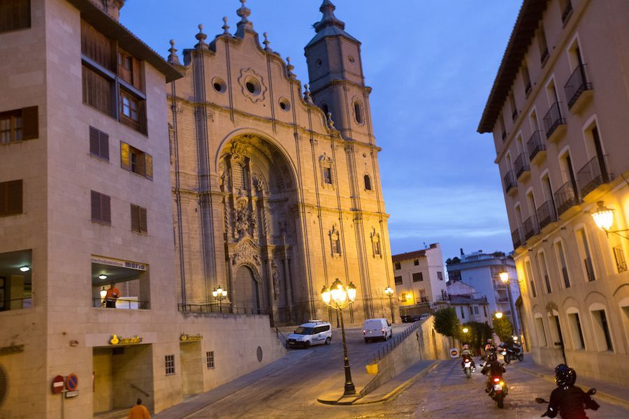 Illuminated cathedral at dusk on a sloping city street, with buildings and a few pedestrians nearby