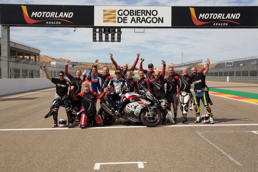 Motorcycle racing team posing on track under MotoLand and Gobierno de Aragón banners, celebrating with raised arms