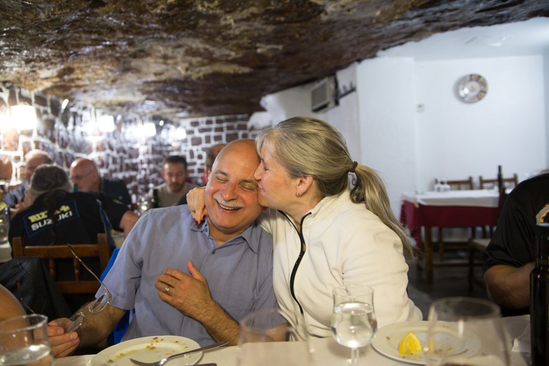 Couple smiling at a table in a rustic restaurant, woman leaning in to kiss the man’s cheek