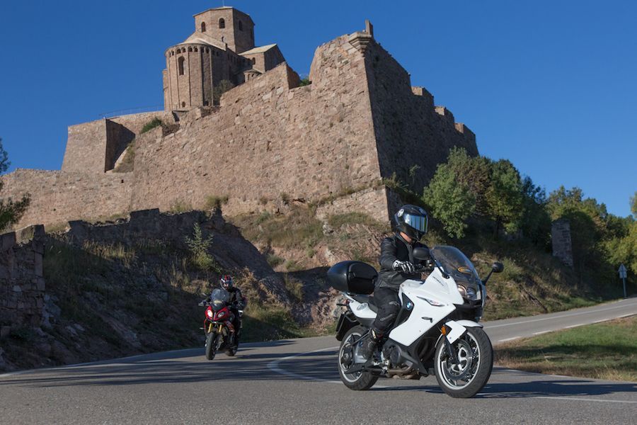Two motorcycles on a road beside a stone castle under a clear blue sky