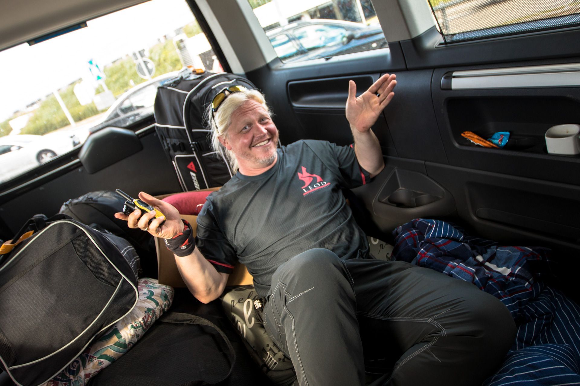 Person smiling in a car’s back seat, holding a phone with one hand and gesturing with the other.