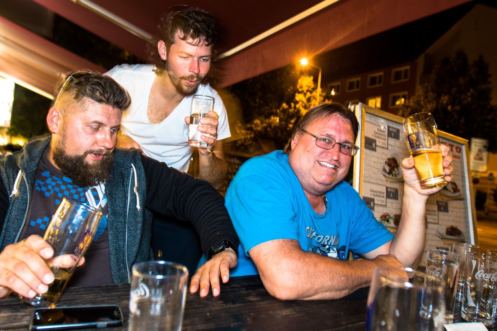 Three men leaning on a bar, smiling and holding drinks in a warmly lit pub.
