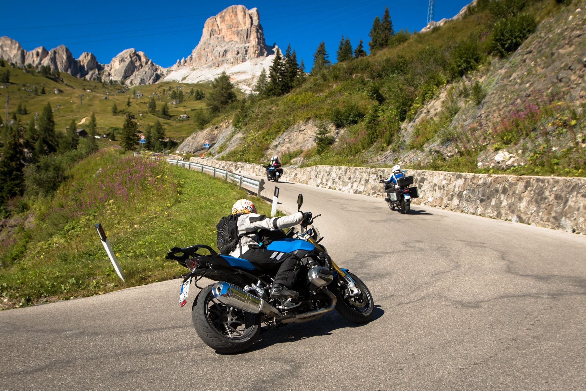 Motorcycles parked on a mountain road with rocky peaks, pine trees, and clear blue sky