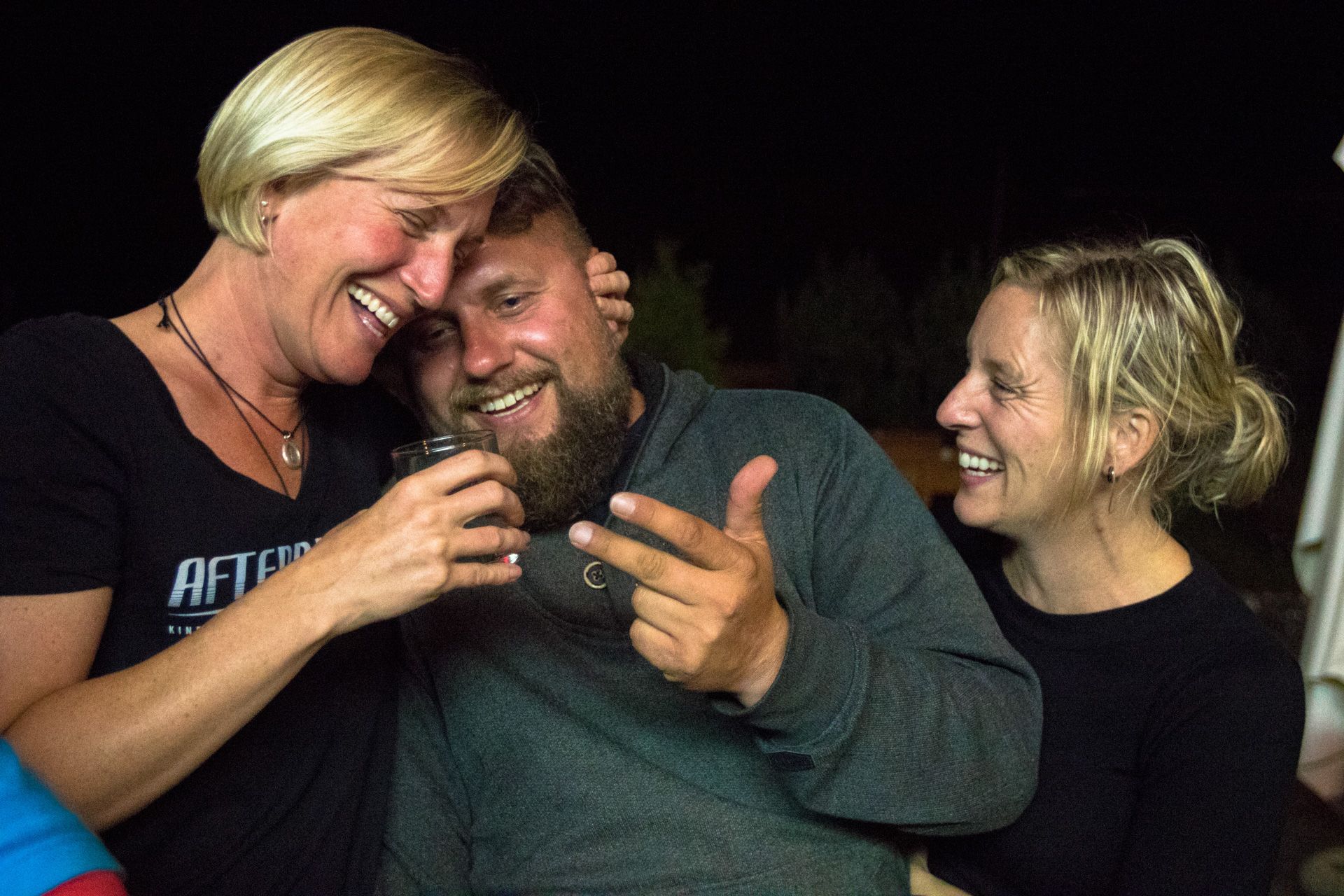 Three smiling people huddle together at night, one holding a cigarette and another a drink.