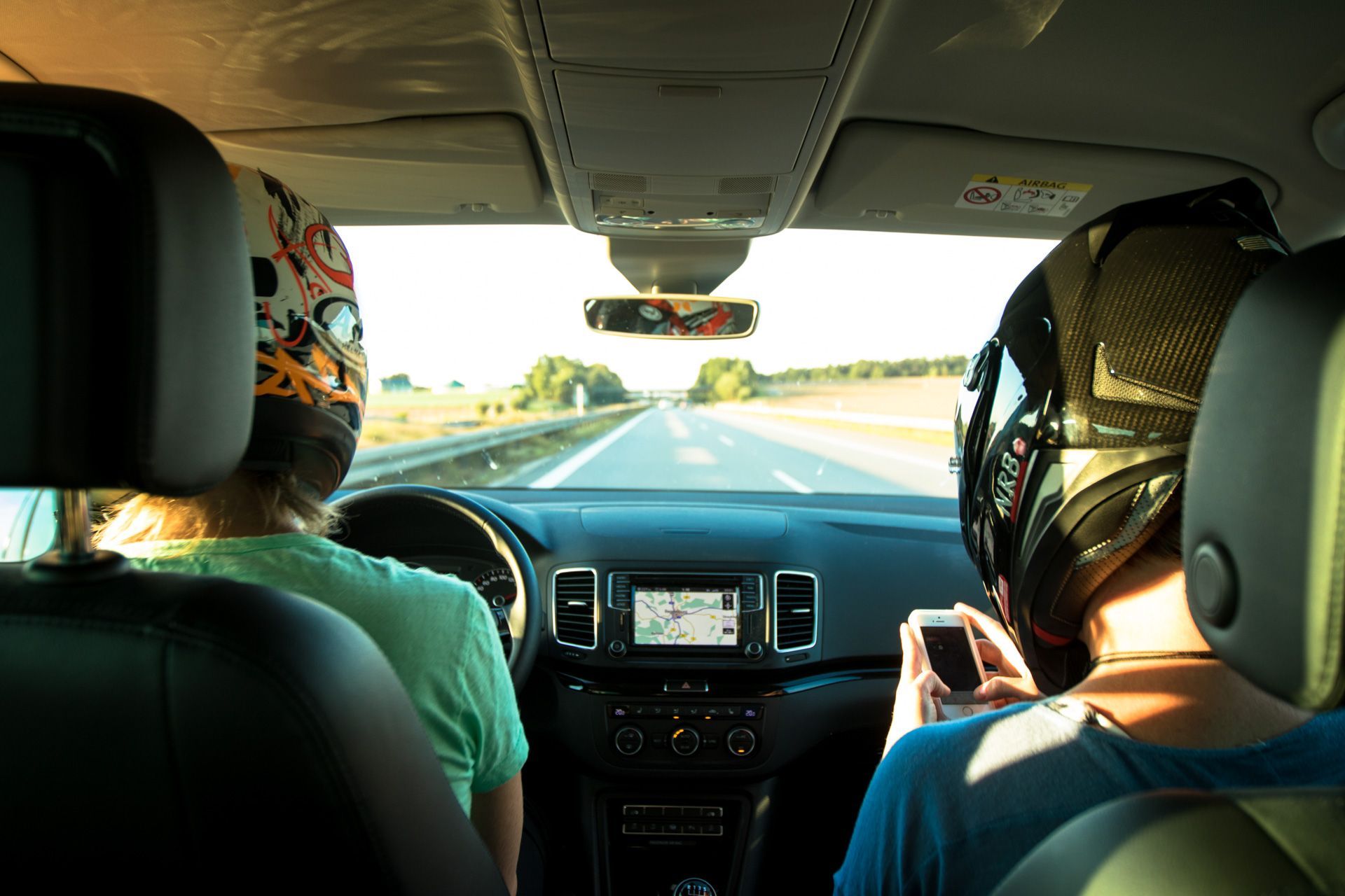 Two helmeted passengers ride in a car, viewed from the back seat on a sunny highway.
