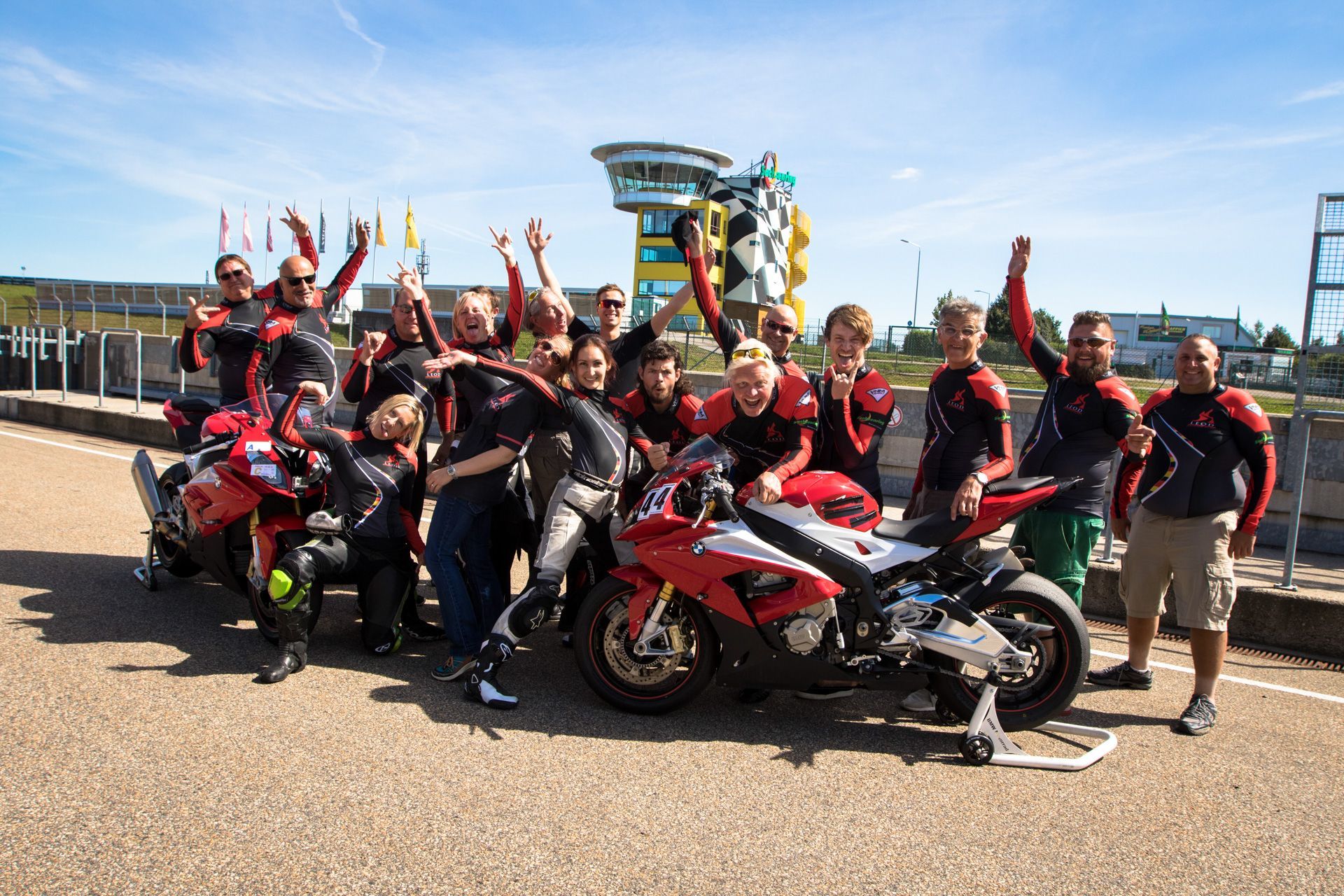 Group of motorcycle racers posing with bikes and raised arms on a sunny trackside area