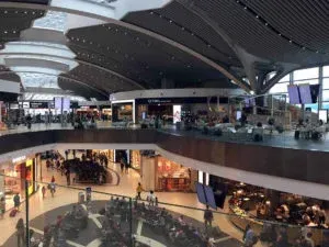 Busy modern shopping mall interior with multiple floors, glass railings, and people walking and sitting