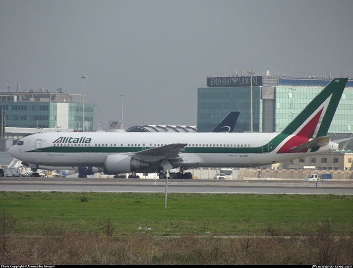 Alitalia passenger jet taxiing on a runway, with city buildings in the background.