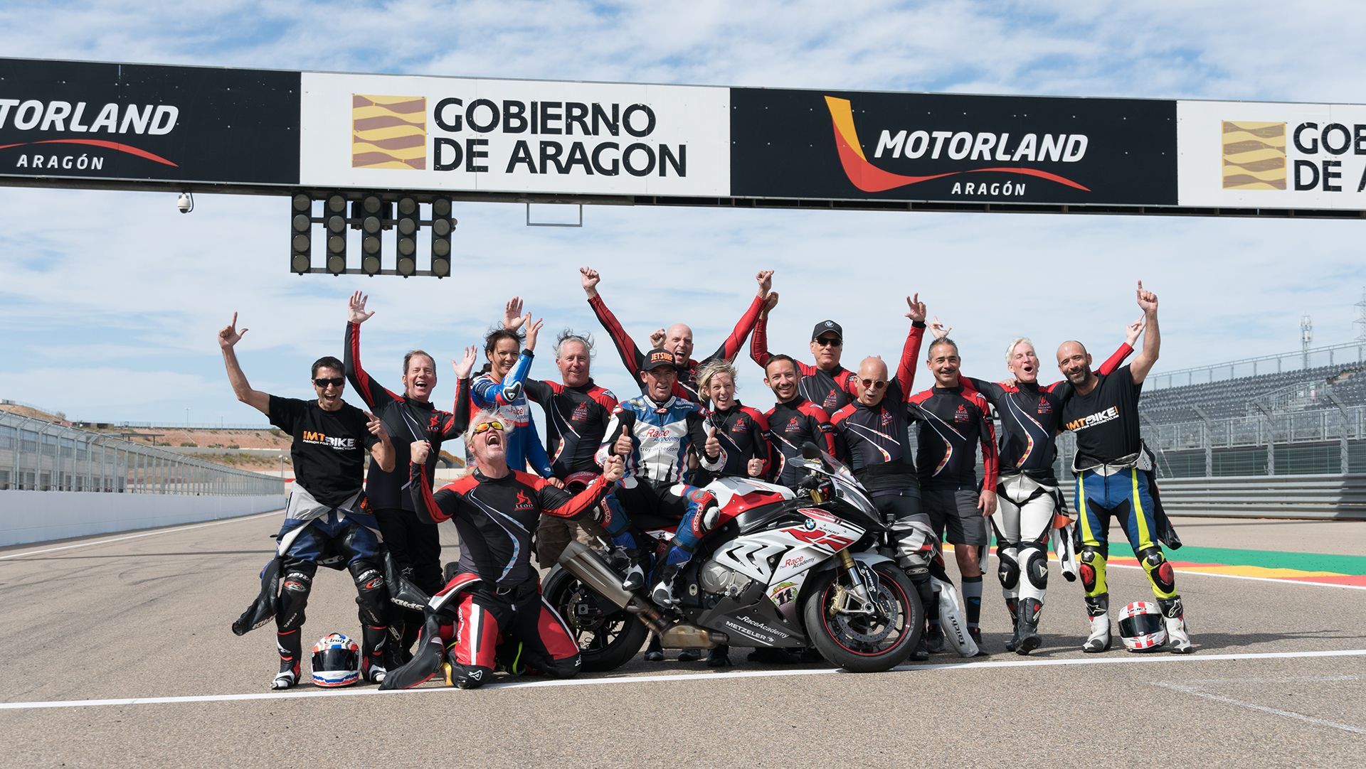 Motorcycle race team posing on the track with trophy, waving in front of sponsor banners.