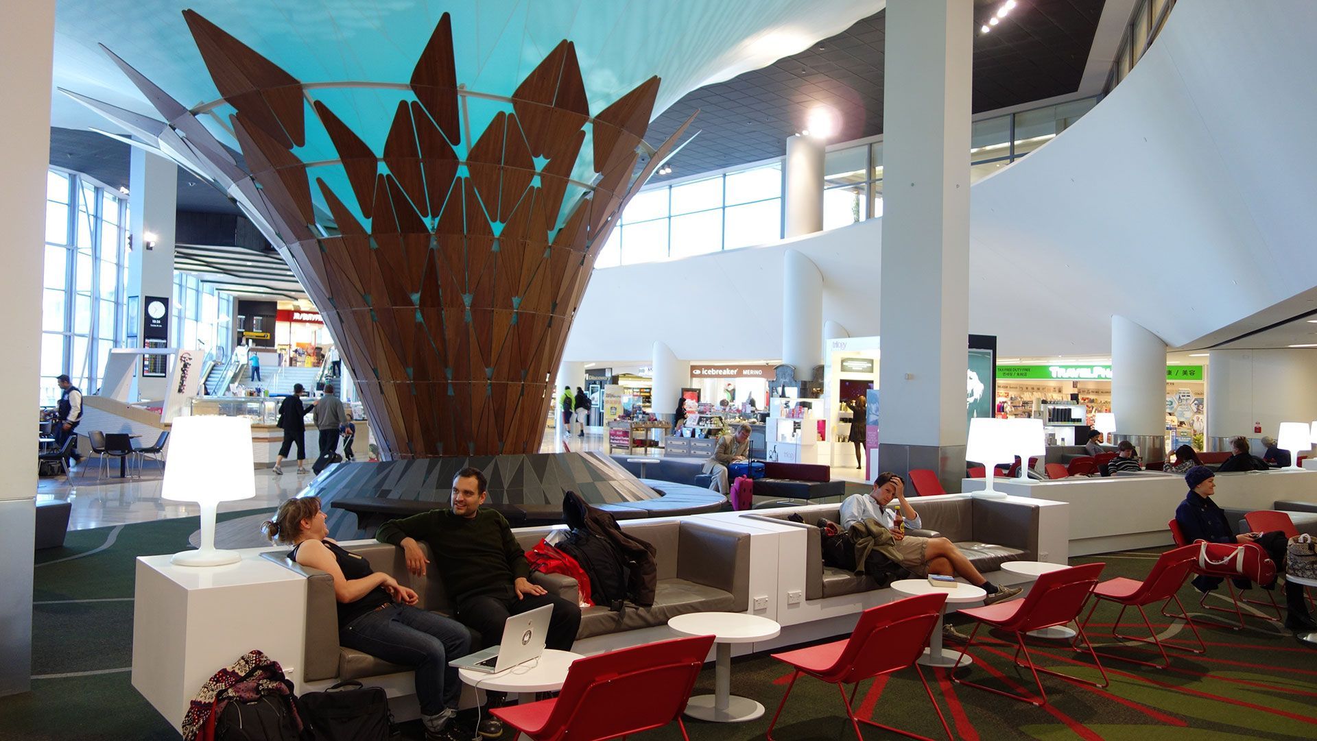 Airport lounge with red chairs, travelers seated, and a large wooden sculpture in the center.