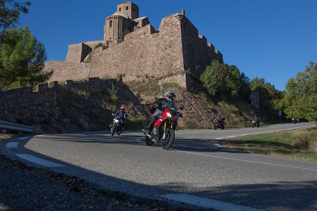 Motorcyclist riding on a winding road beside a stone castle on a hill under a clear blue sky