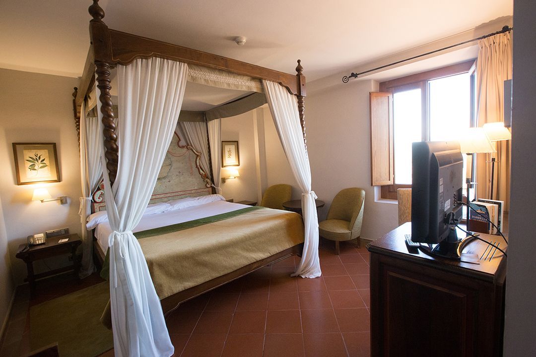 Elegant bedroom with a canopy bed, white curtains, warm lighting, and a dresser by a window.