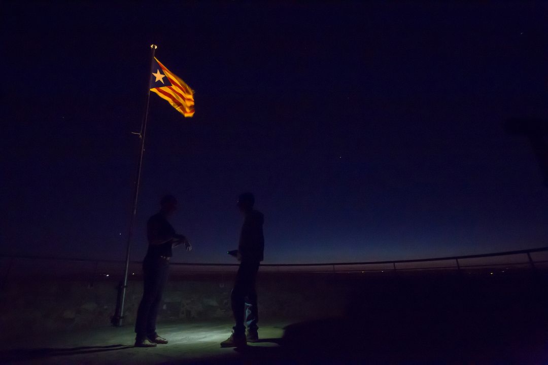 Two silhouetted people stand by a flagpole at night, with an illuminated flag against a dark horizon.