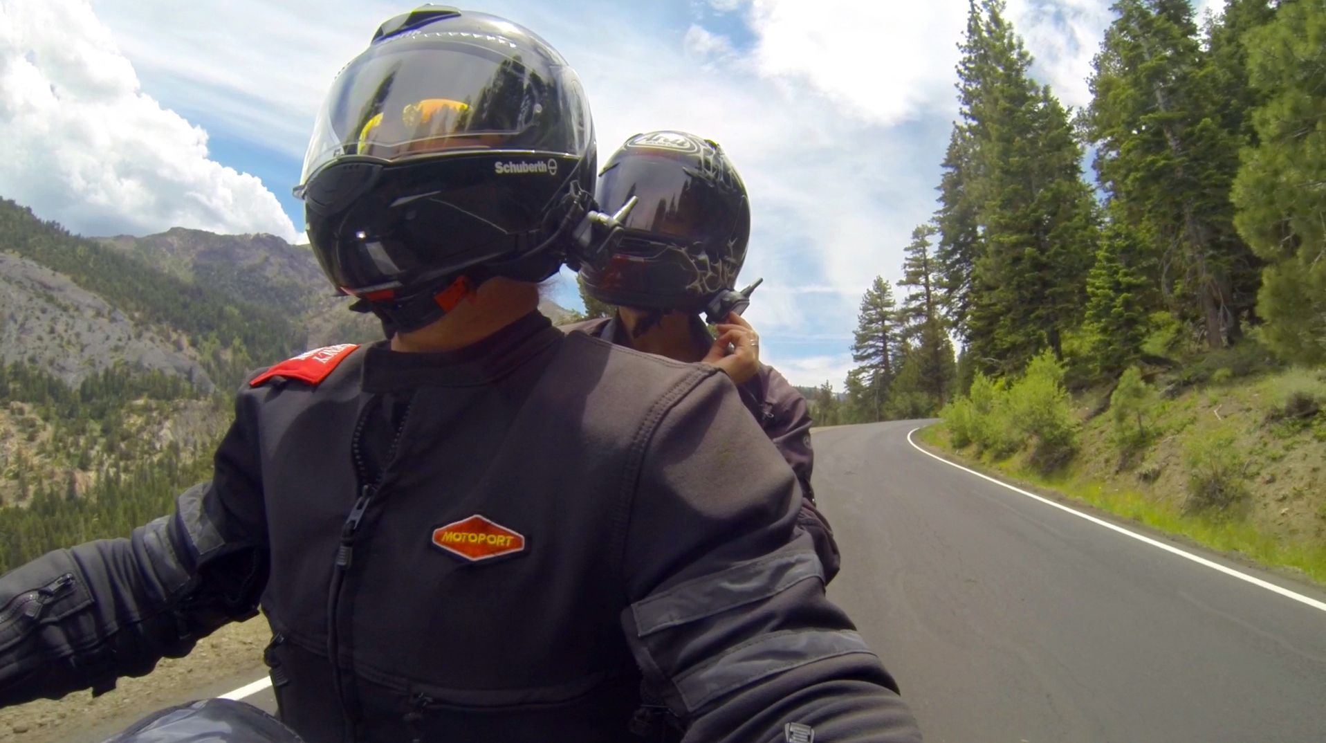 Two motorcyclists in black gear ride on a curving mountain road beside forest and rocks.