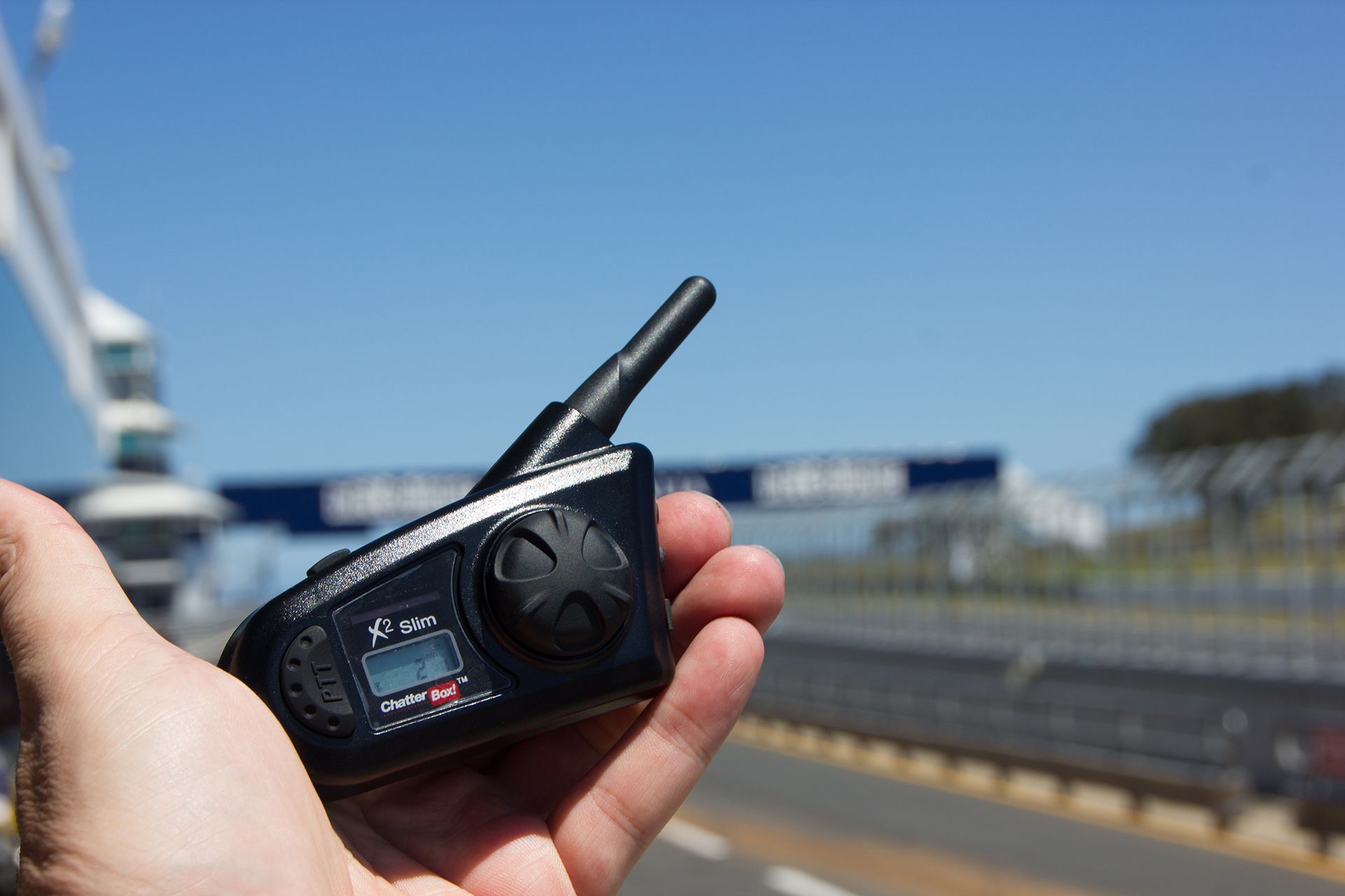 Hand holding a black walkie-talkie with antennas near a racetrack under a blue sky