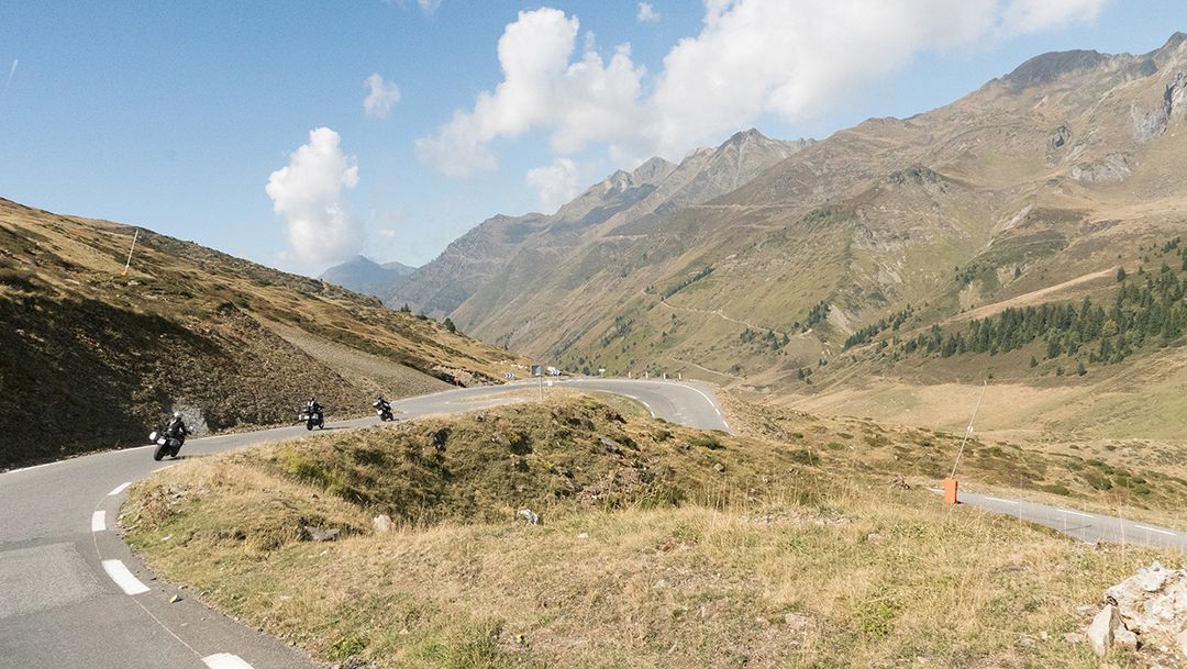 Mountain road winding through dry hills under a blue sky, with a motorcyclist on the left and mountains ahead.