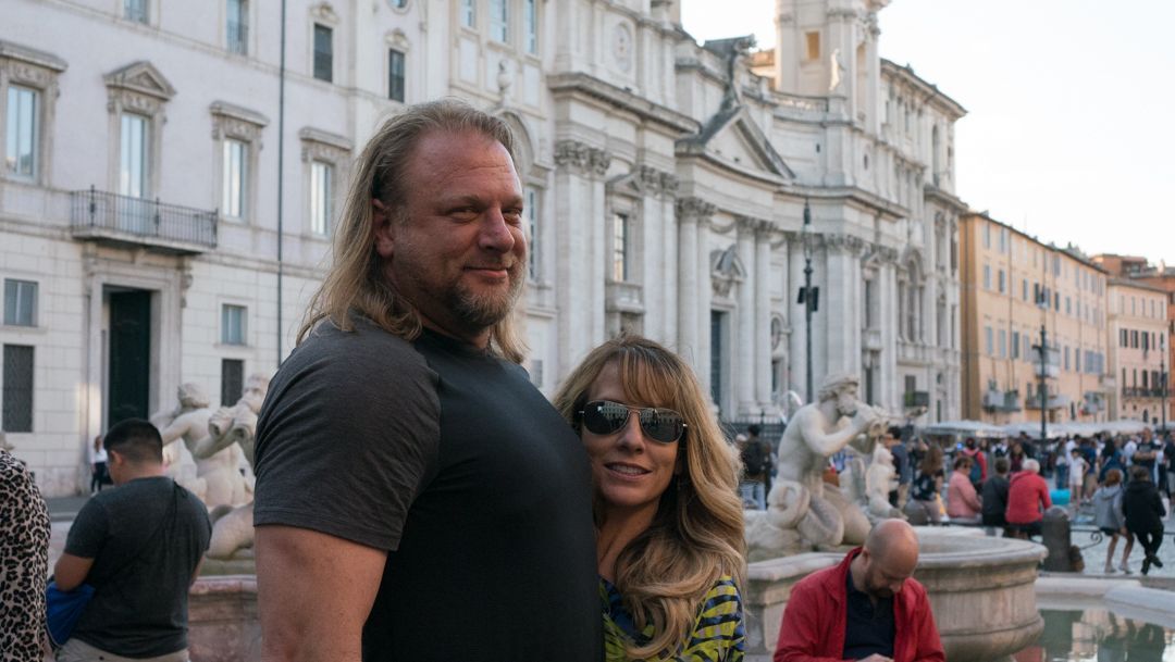 Two people posing by a canal in front of white historic buildings on a crowded waterfront