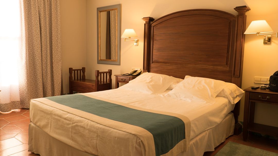 Warm hotel room with a large wooden bed, cream bedding, bedside lamps, and soft curtains.