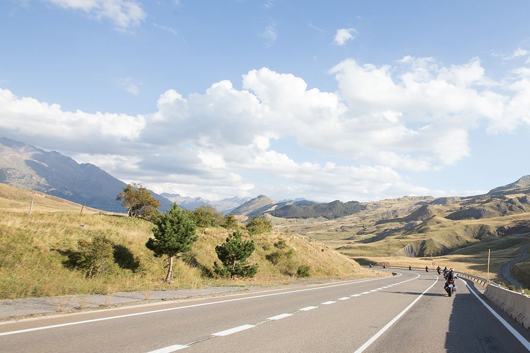 Mountain road with cyclists under a cloudy sky, surrounded by dry hills and distant mountains