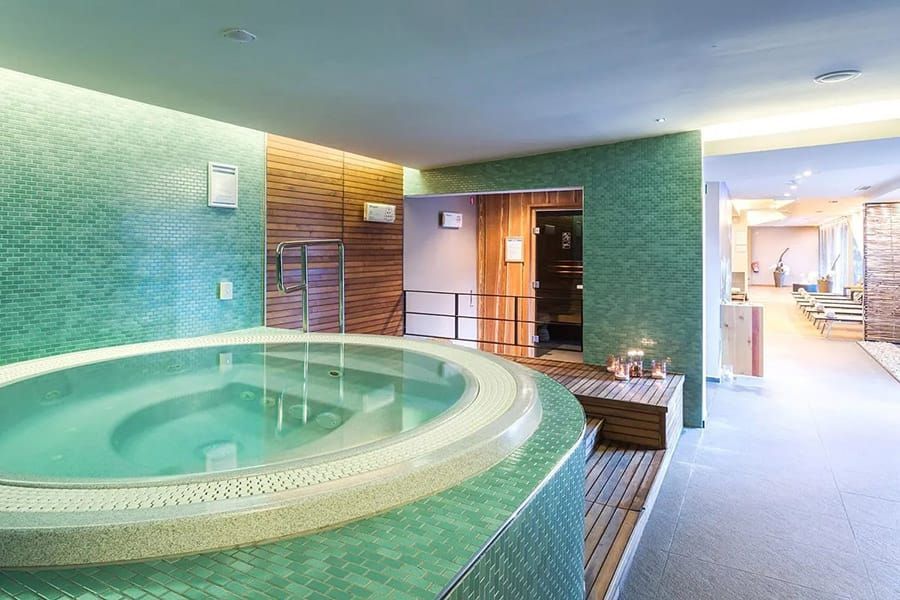 Indoor spa with a round tiled hot tub, green walls, wood accents, and lounge seating nearby
