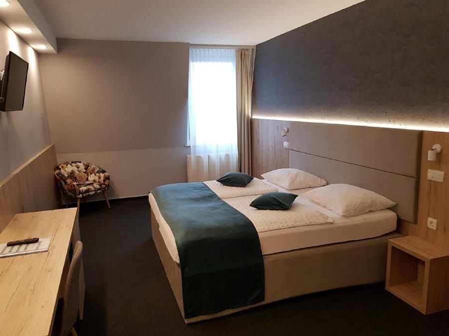 Modern hotel room with twin bed, teal accents, desk, chair, and window blinds