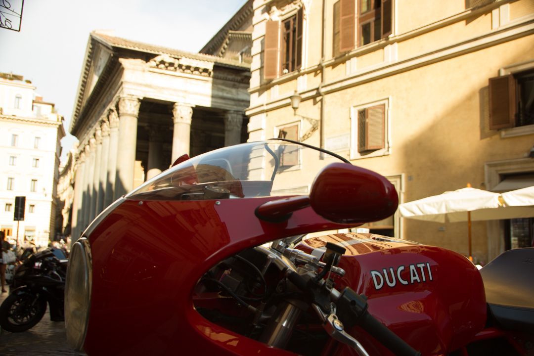 Red Ducati motorcycle parked on a sunlit street with historic buildings in the background