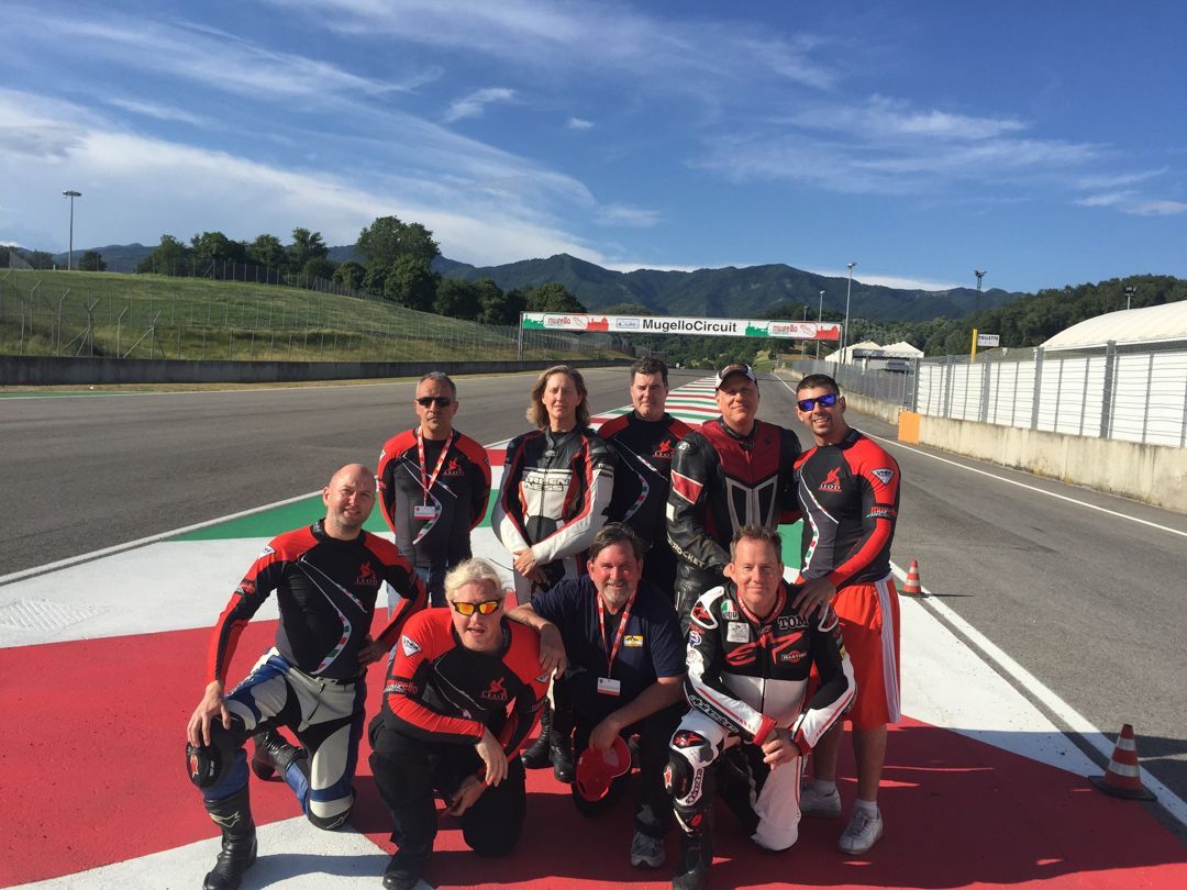 Nine race car drivers in red-and-black suits pose on a racetrack with a bridge in the background