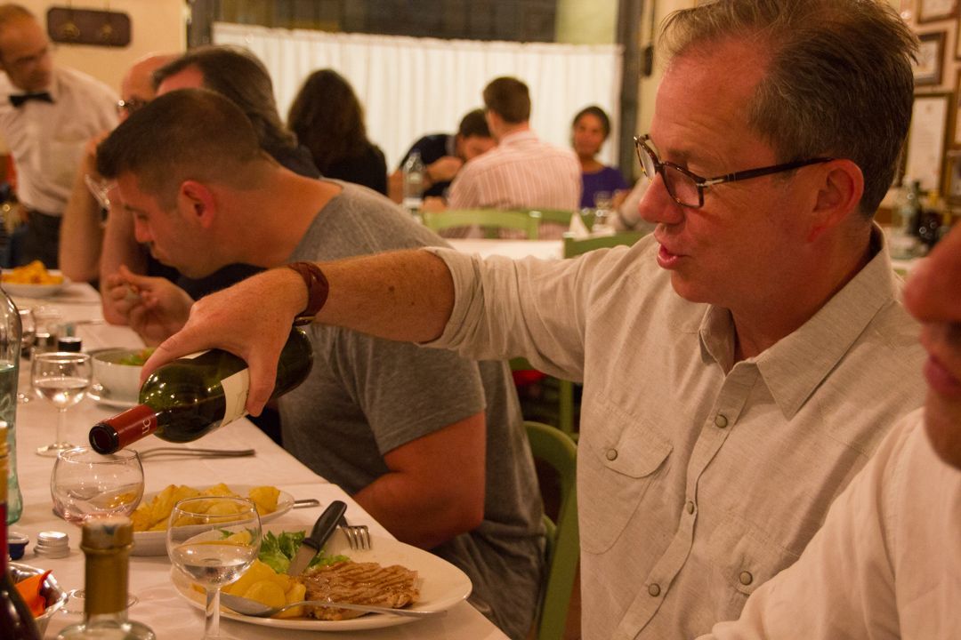 Man pouring red wine at a dinner table in a restaurant with plates, glasses, and other diners nearby