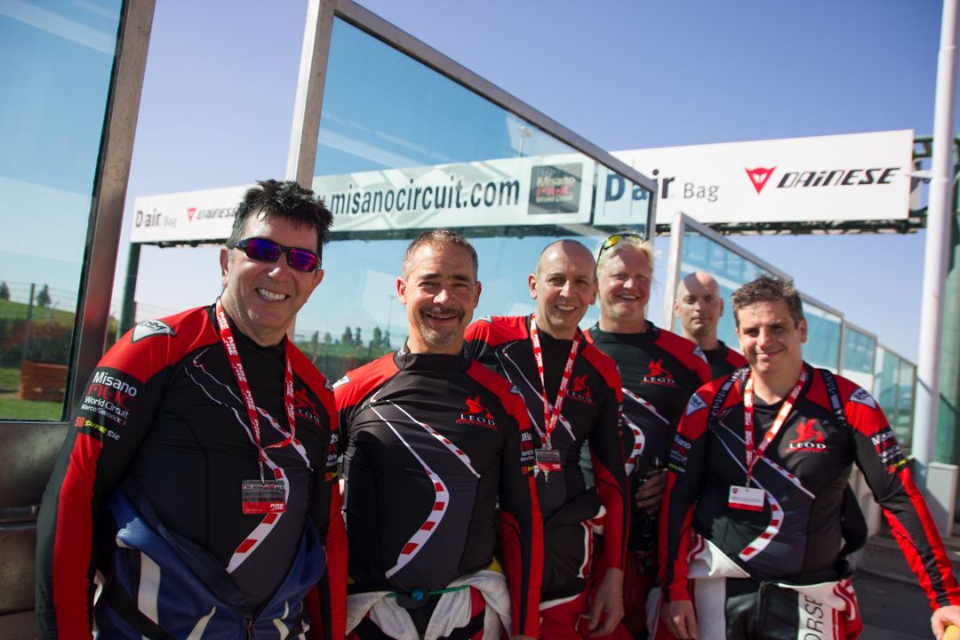 Four cyclists in matching black and red jerseys posing outdoors at a race finish banner