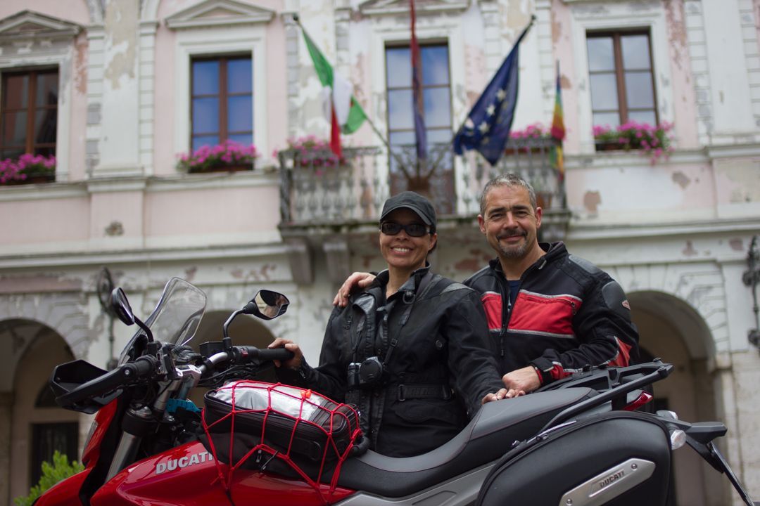 Two people on a scooter in front of a building with flags and pink flowers