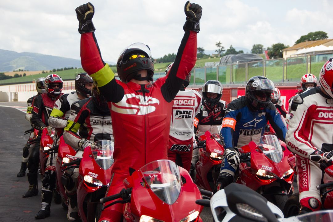 Motorcycle racers on red bikes line up at a track; one rider in red raises both fists in celebration.
