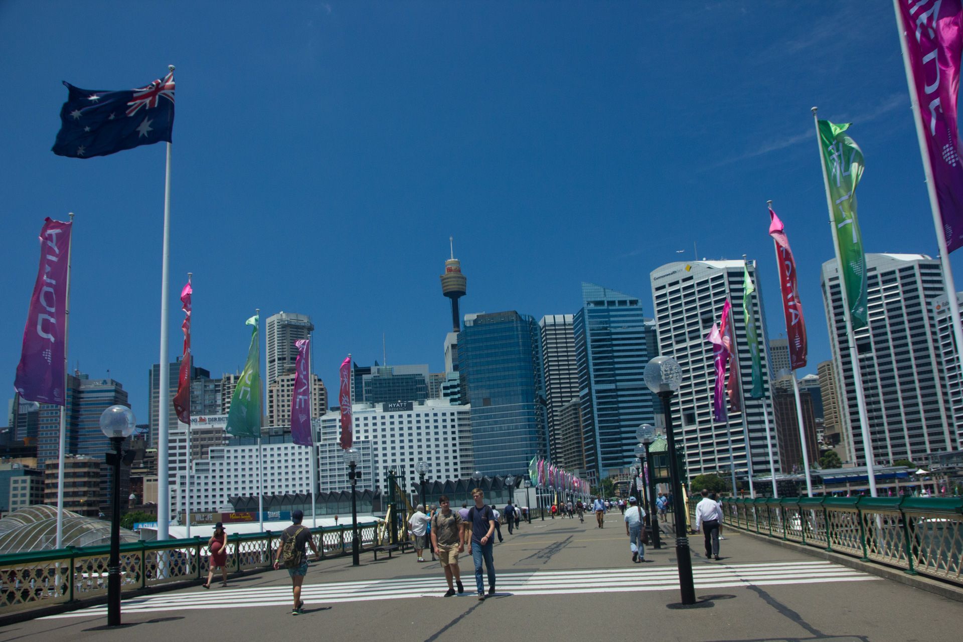 Pedestrian bridge leading to a modern city skyline with colorful flags under a clear blue sky