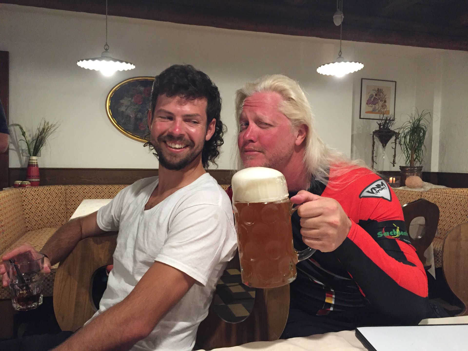 Two men in a pub smiling and holding large foamy beer mugs at a table