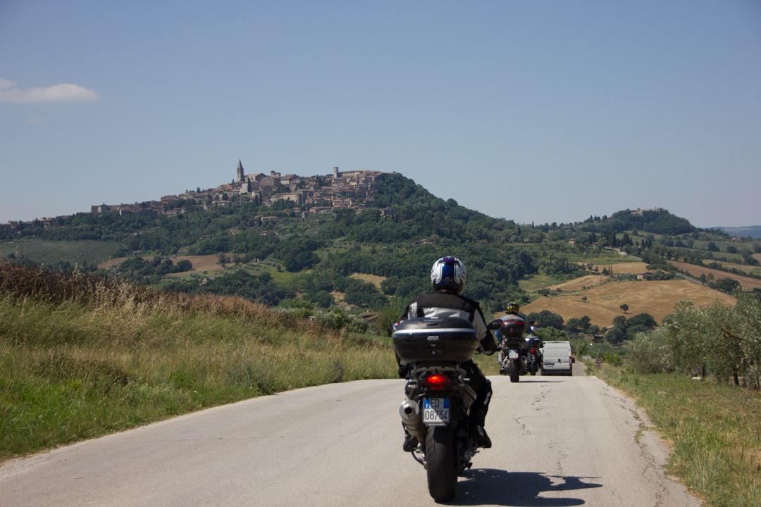 Motorcyclists riding along a rural road toward a hilltop village under a clear sky