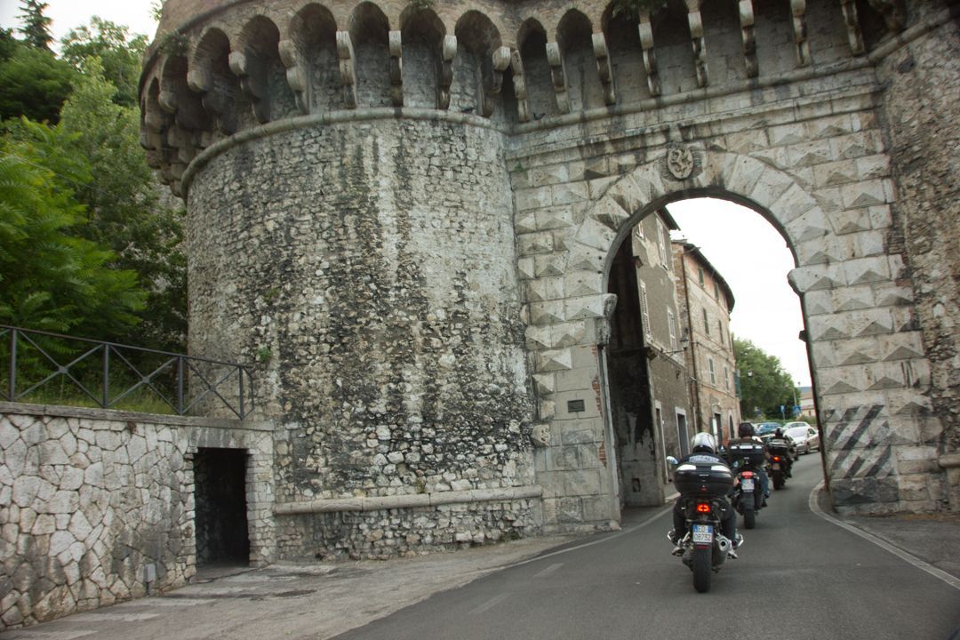 Motorcyclists pass through a stone city gate with an arched opening and weathered walls.