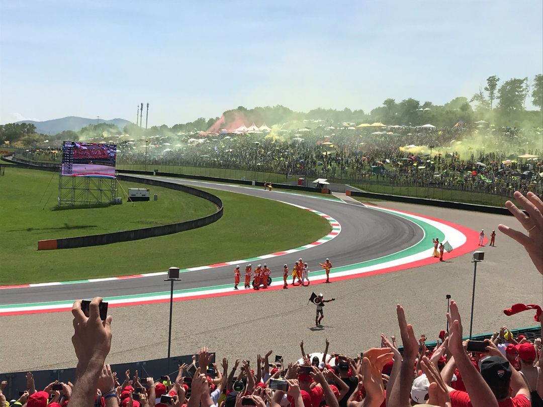 Race cars speeding through a left-hand bend on a packed track, with cheering crowd in the foreground.