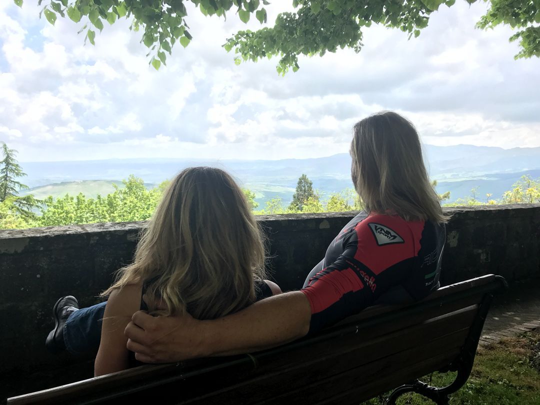 Two people sit on a bench overlooking a cloudy mountain landscape and green valley.