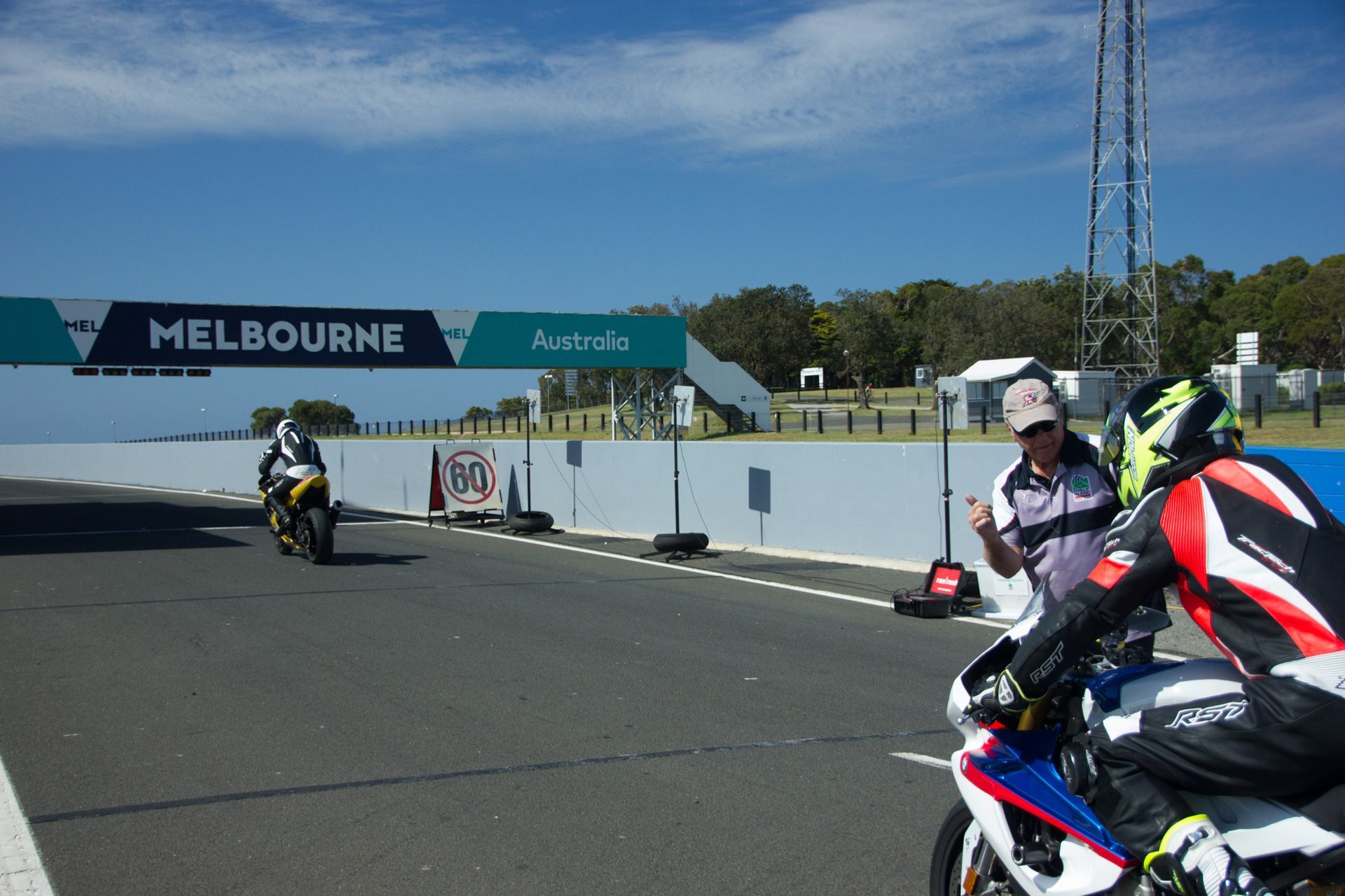 Motorcyclists on a racetrack near a Melbourne sign, with one rider in the foreground and others ahead.